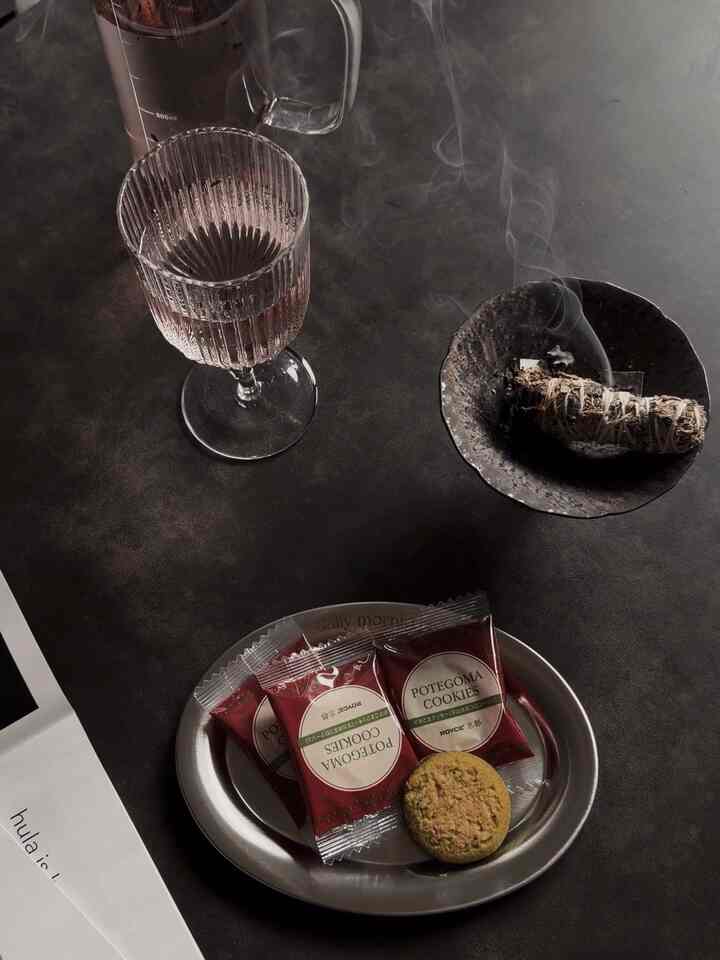 Cozy space featuring a dark gray toned table with a ribbed glass, incense dish emitting smoke, and a plate of cookies