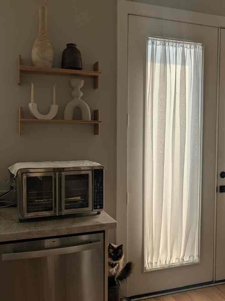 White-toned kitchen featuring sculptural vases on wooden wall shelves and sheer white curtains on the balcony door, creating a cozy atmosphere