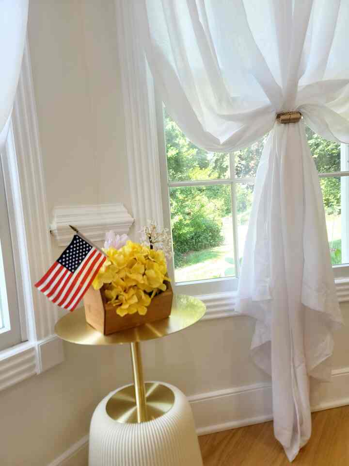 Sunlit veranda corner with sheer white curtains, round gold side table featuring bright yellow faux flowers and a small American flag