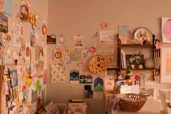 Warm beige-toned small vintage room featuring a wall covered with various postcards and posters, a wooden shelf with vintage cassette player creating a cozy workspace