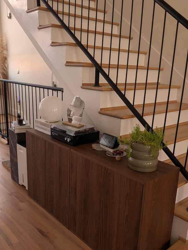 Wood tone modern living room entrance featuring storage cabinet with record player, lamp, and plant under staircase