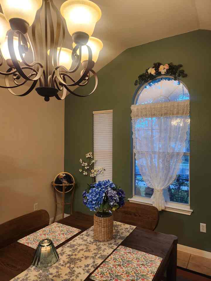 Dining room with olive green walls and arched window featuring lace curtains, wooden dining table and floral decor in a romantic setting