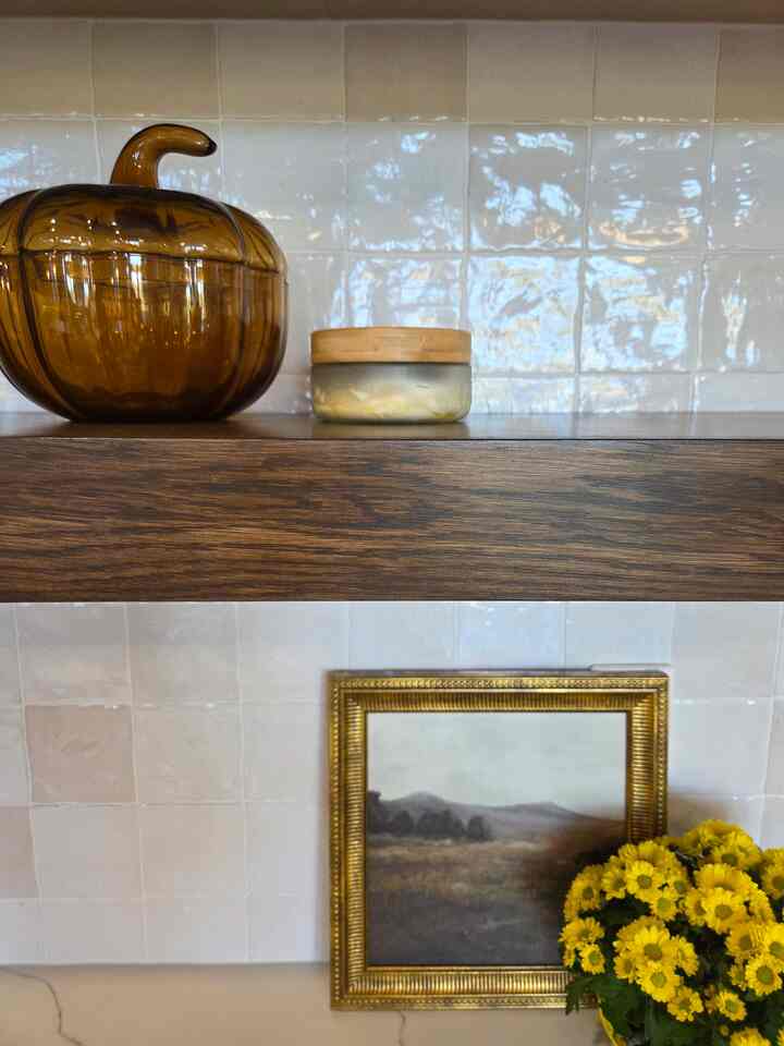 Cozy interior space featuring wood-toned shelf with amber pumpkin-shaped glass bowl and small container above, golden framed painting and yellow flowers below