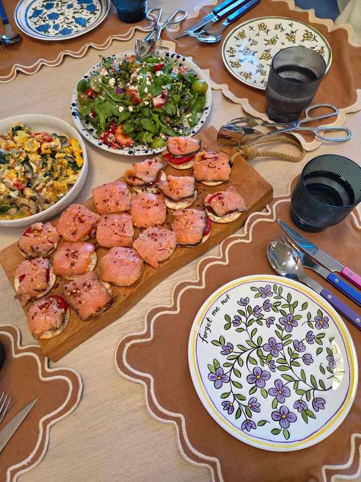 Warm wood tone dining table set with beige placemats, floral plates, salad, and smoked salmon crackers for a cozy home party