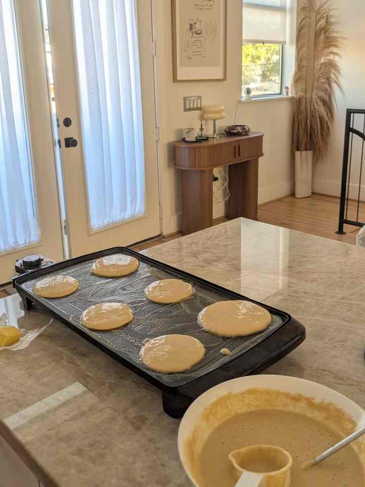 A natural-toned kitchen featuring a marble dining table with an electric griddle cooking pancakes in a white and brown color scheme