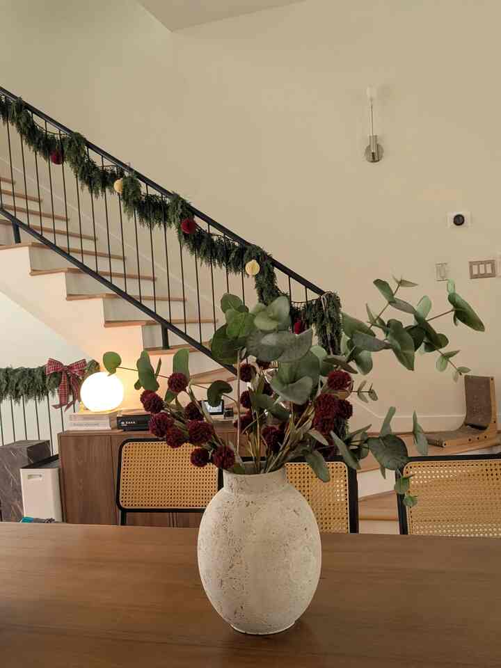 Natural color dining room featuring a wooden dining table, rattan chairs, and a large ceramic vase with plants at center, cozy atmosphere
