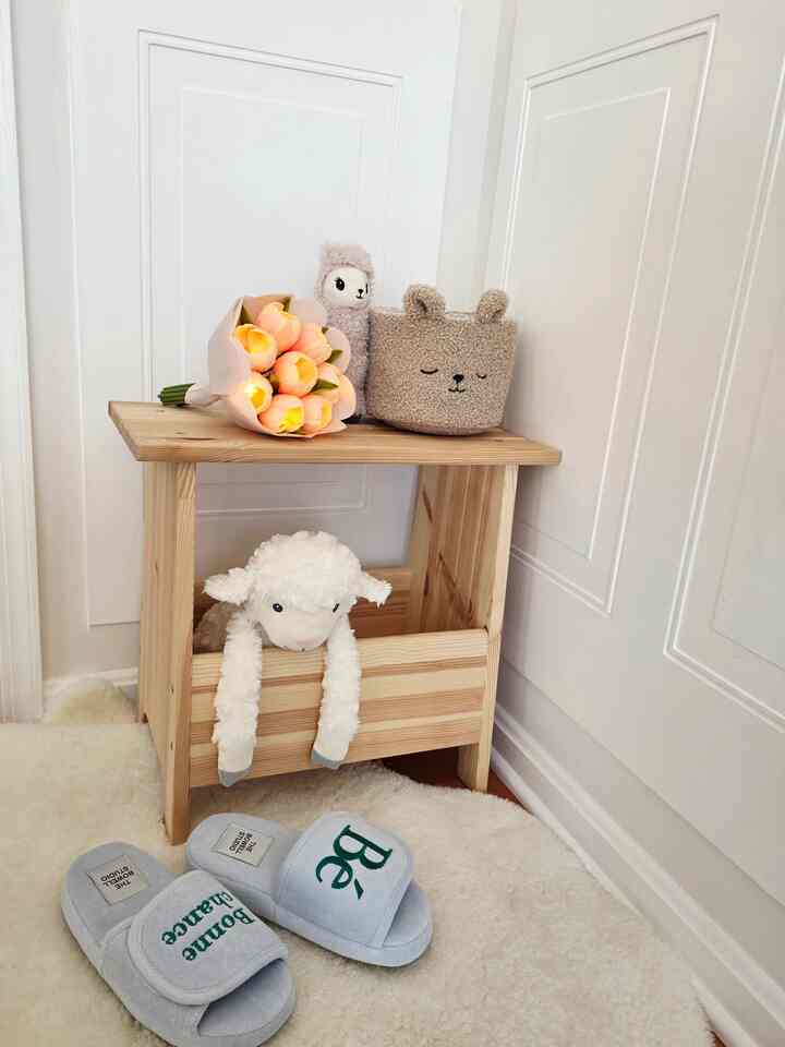 White and wood tone kids' room corner featuring tulip mood lamp, plush toys on wooden stool, soft rug and cozy slippers for a warm atmosphere