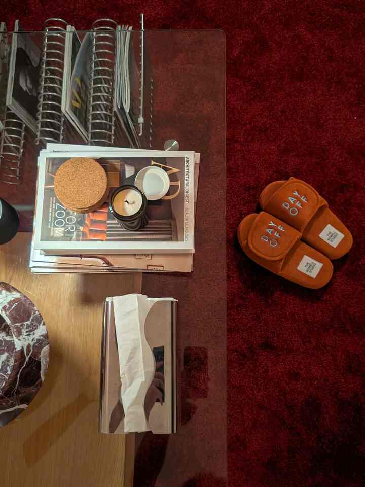 Living room space with dark red rug featuring round brown slippers and gold metal magazine holder on wooden and glass table with magazines and tissues