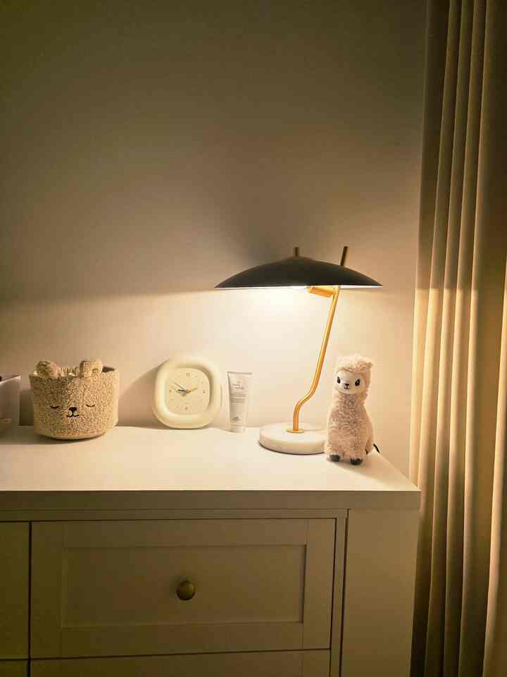 Cozy bedroom corner with warm lighting featuring a gold table lamp and soft toys placed on a white nightstand