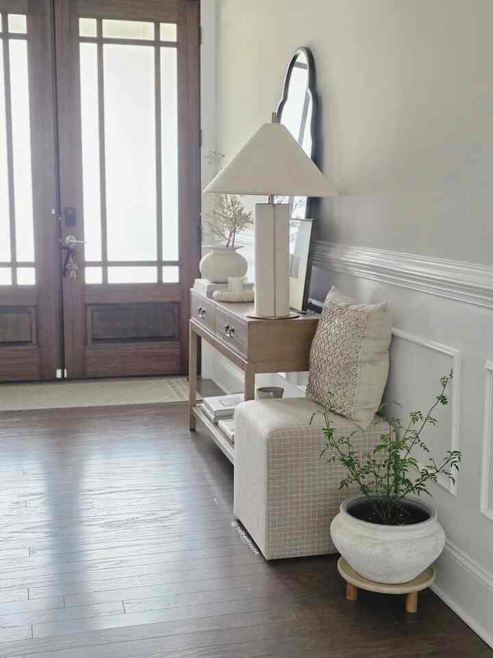 Bright white and brown-toned entrance featuring a console table with lamp, ottoman with cushion, and a plant in a natural styled space