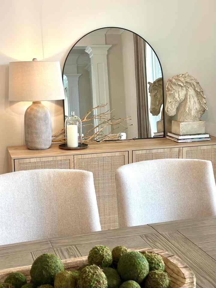 A natural-toned dining room featuring beige table lamp, large mirror, and horse head sculpture on a wooden sideboard with moss balls on the table.