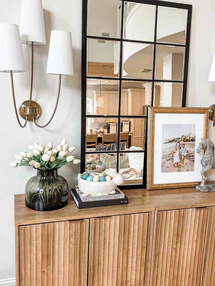Natural brown wood tone entrance featuring large grid mirror, plant stand with white tulips, wall sconces, and framed photo for cozy decor