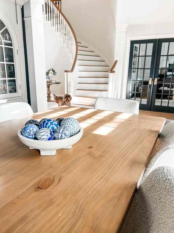 A bright dining room with white walls and wood-tone dining table, featuring blue and white ceramic orbs centerpiece and a dog near the staircase, creating a cozy atmosphere
