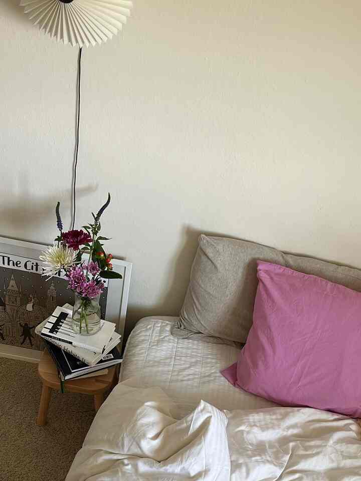 A cozy bedroom in white and beige tones featuring a bed with pink and gray pillows and a side table adorned with a vase of flowers