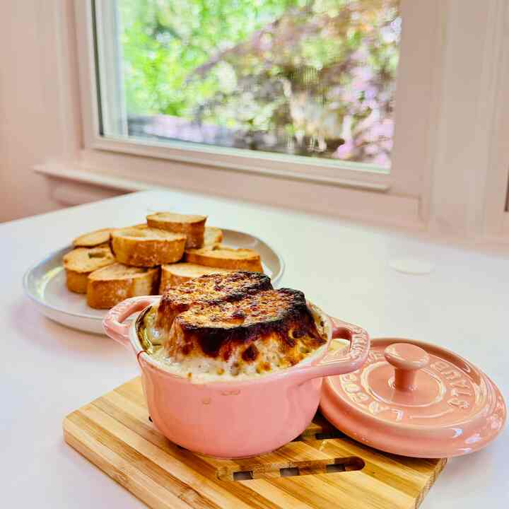 Bright orange Staub pot with toasted bread on white kitchen table by a window showing green trees, creating a warm atmosphere