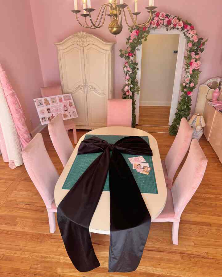 Pink dining room featuring a white oval table with six pink chairs and a large black velvet bow centerpiece, accented by a vintage wardrobe and floral-decorated mirror