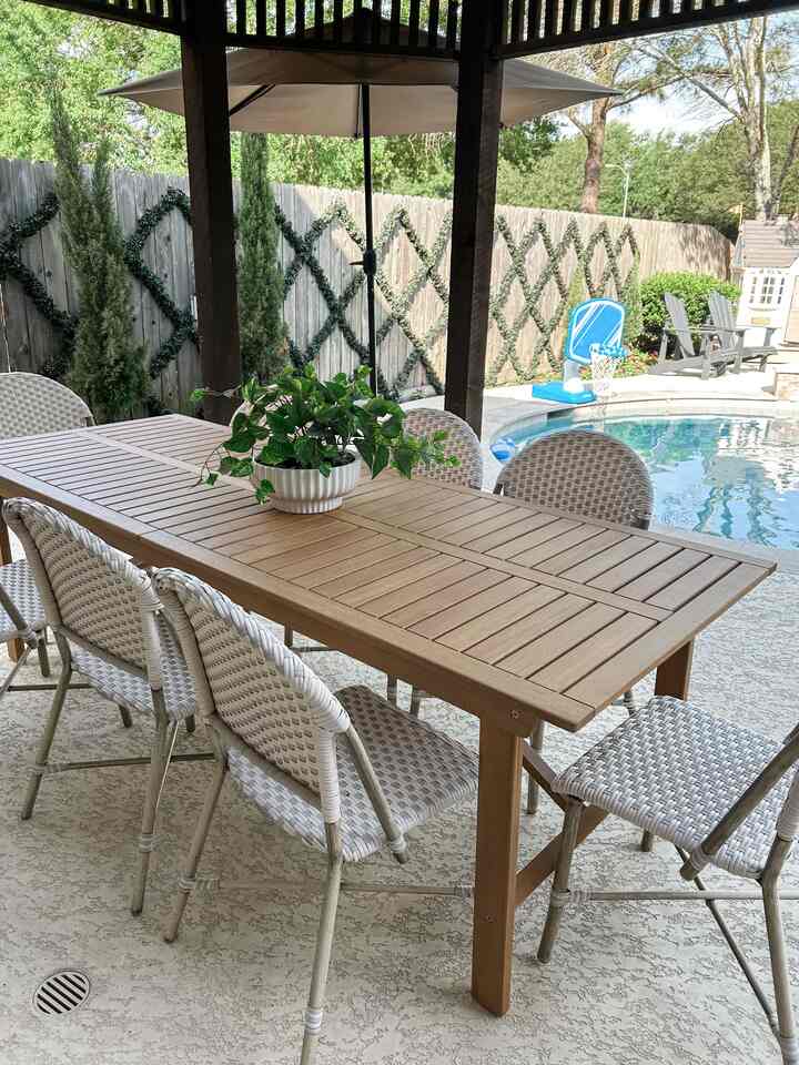 Natural tone outdoor balcony space featuring wooden dining table and outdoor chairs with green plants in the background, creating a relaxed atmosphere