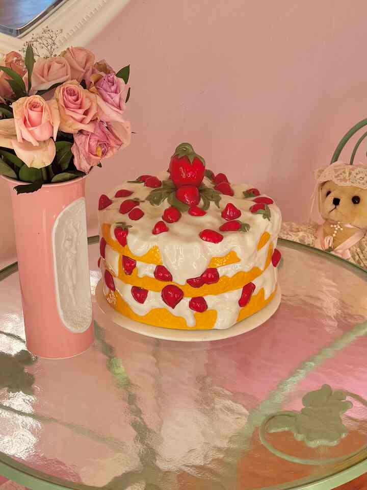 A soft pink-toned dining room featuring a glass dining table with a vintage strawberry shortcake ceramic dish and pink vase with flowers, accented by a decorative teddy bear on the right, creating a charming atmosphere