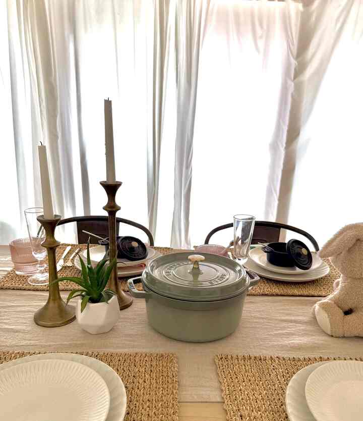A warm, natural-toned dining room featuring a Staub pot and bronze candle holders centered on a cozy dining table