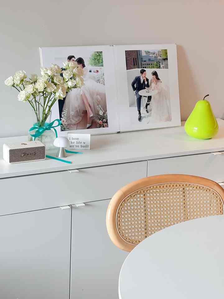 White and natural toned apartment dining room featuring a round dining table, vase with flowers, and a wedding photo book creating a cozy ambiance