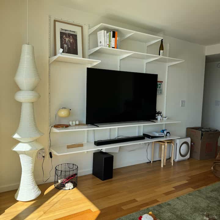 Modern living room in white and wood tones featuring wall-mounted shelves, a large TV, and sculptural floor lamp