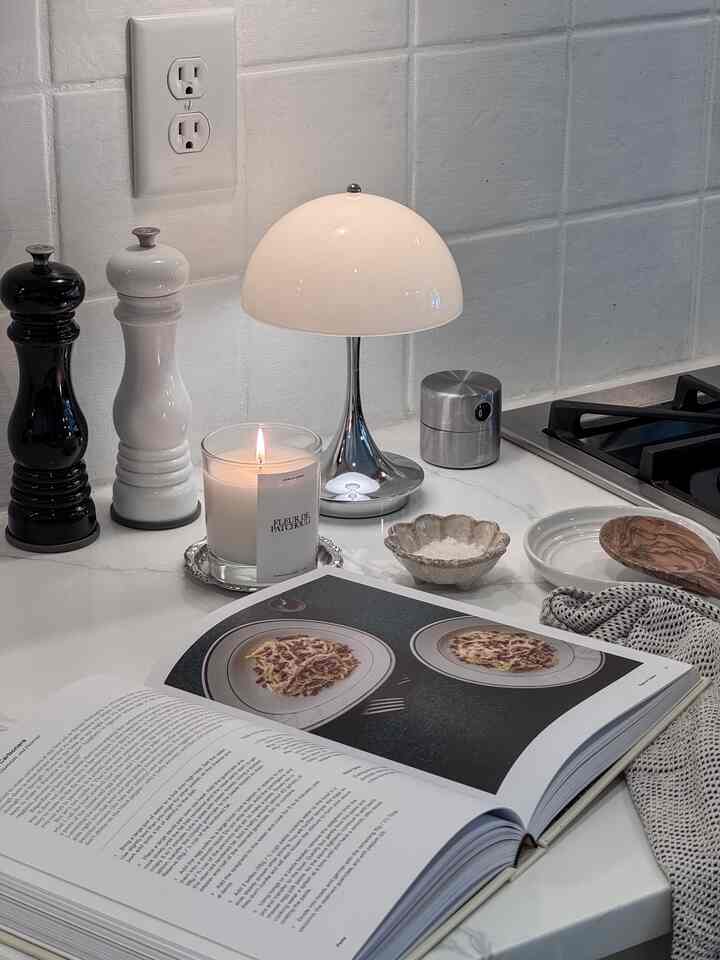 Modern kitchen space with white tiled wall and countertop featuring Louis Poulsen's Panthella table lamp, lit candle, and an open cookbook