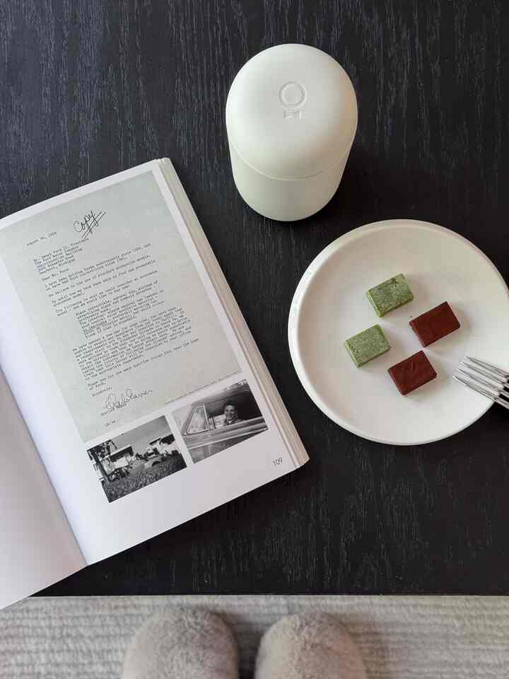 Dark wood tone table featuring a white mug, plate with desserts, and an open book creating a tidy home cafe scene