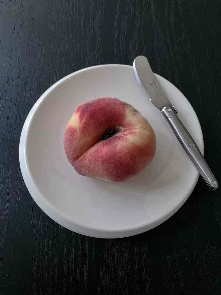 A simple setting on a dark wood tone table featuring a flat peach on a white plate with a cheese knife placed on the right