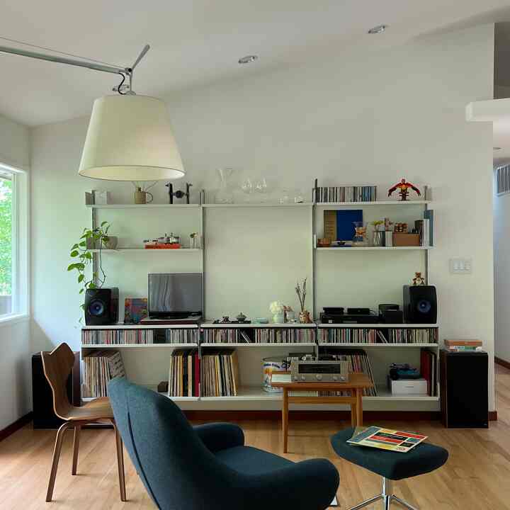 Nordic style living room with white walls and wood flooring, featuring a blue armchair and ottoman center front, large shelving unit and Artemide floor lamp on wall, neat and modern