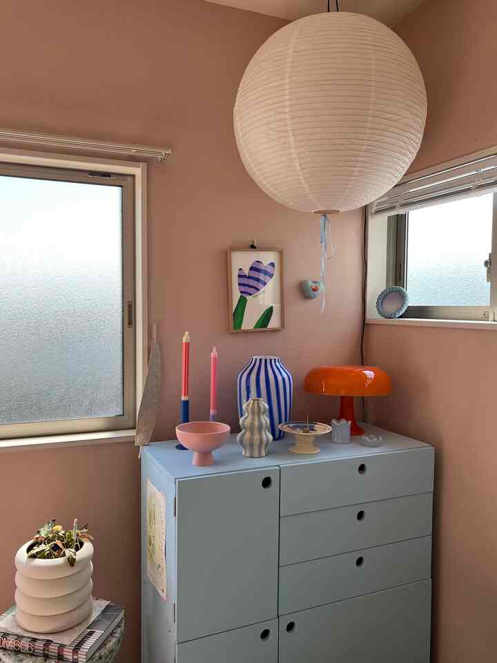 ベージュとライトブルーの色調の部屋、窓際にライトブルーの収納棚があり、オレンジ色のテーブルランプと様々な小物が配置された雰囲気のあるインテリア
