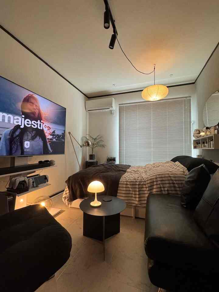 ナチュラルモダンのトーンでまとめられた狭い寝室、ブラインド付きの窓とブラックソファ、テーブルランプが配置された雰囲気のある空間