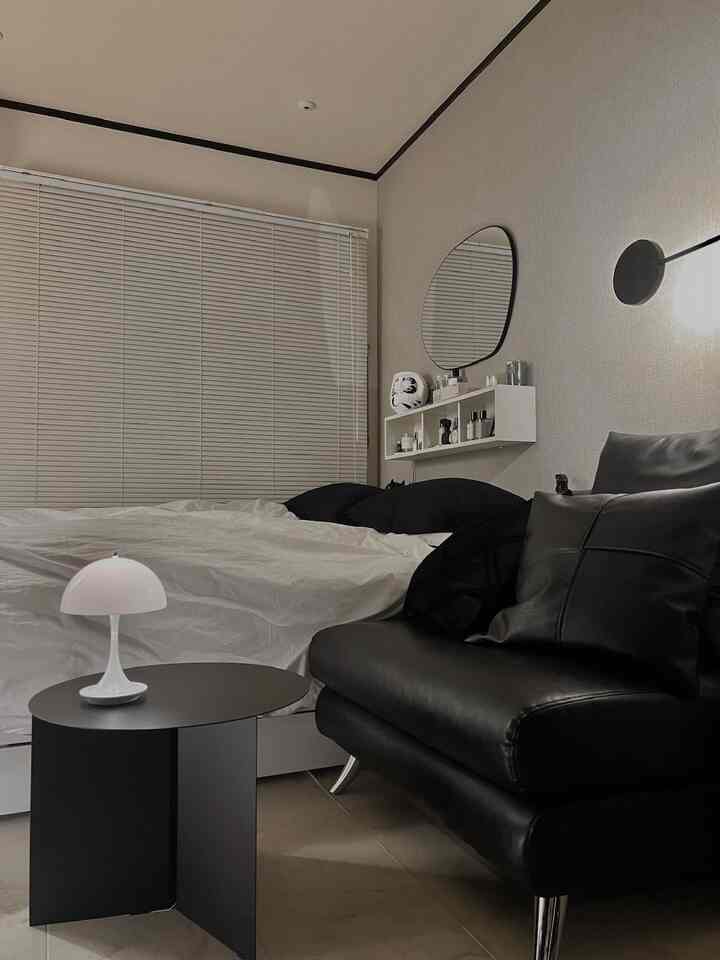 ナチュラルベージュの寝室兼リビング、黒い革張りソファと白いベッド、モダンな照明とシンプルなブラインドが配置された落ち着いた空間