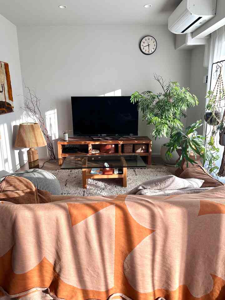 明るいホワイトのリビング、ナチュラルな木目調家具と植物が配置され、オレンジの柄のブランケットとテーブルランプが温かみのある印象的な空間を演出