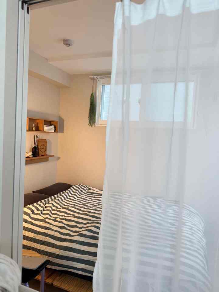 明るい白とベージュのトーンの狭い寝室、中央右にベッドと壁掛け棚が配置されたシンプルな空間