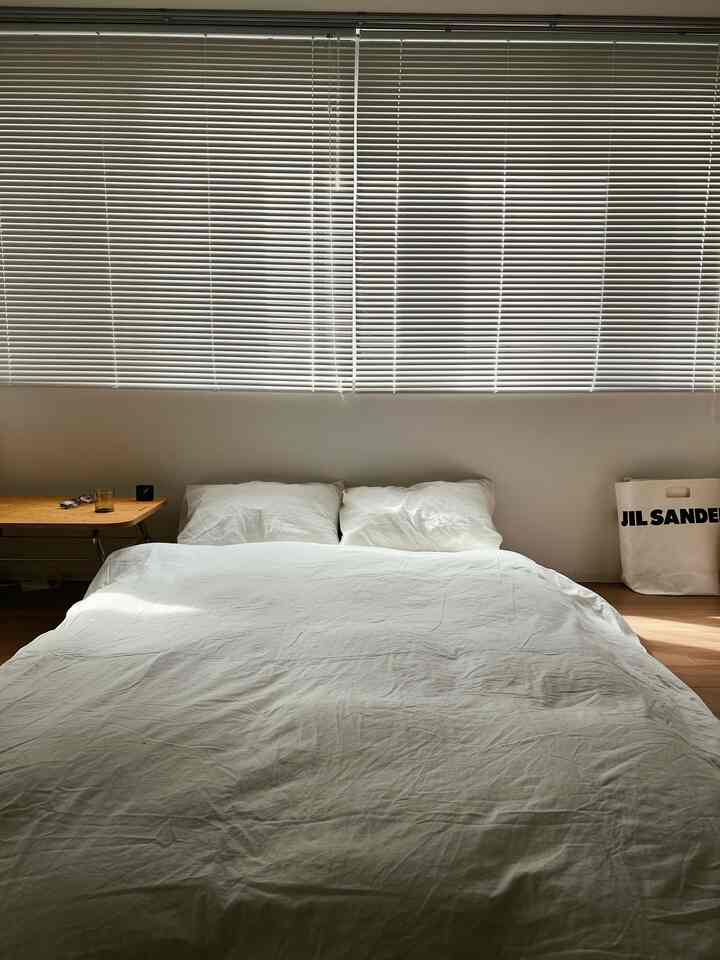 明るいホワイトトーンの寝室、中央に白いベッドと寝具、左側に木製サイドテーブルと窓のブラインドがあるシンプルな空間