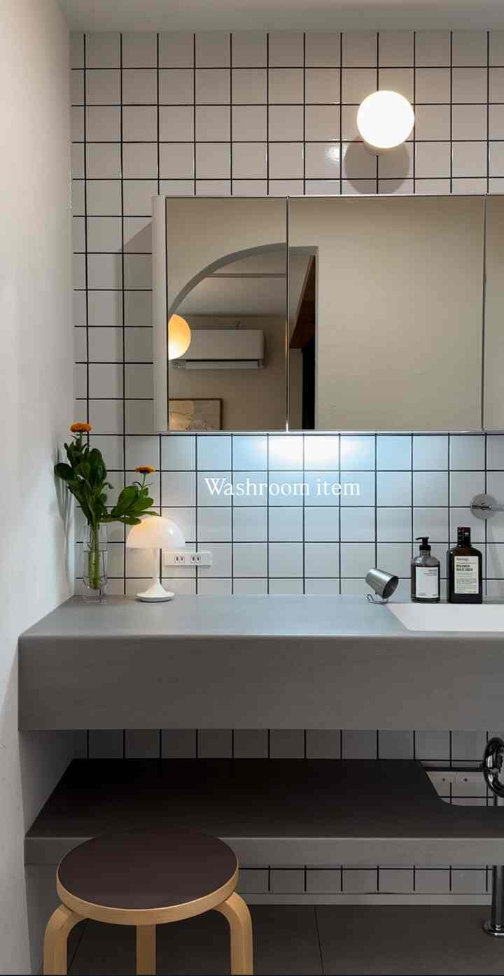白いタイル壁とグレーのシンクカウンターがあるモダンなバスルーム、木製脚のスツールと洗練されたアクセサリーが配置されたインテリア