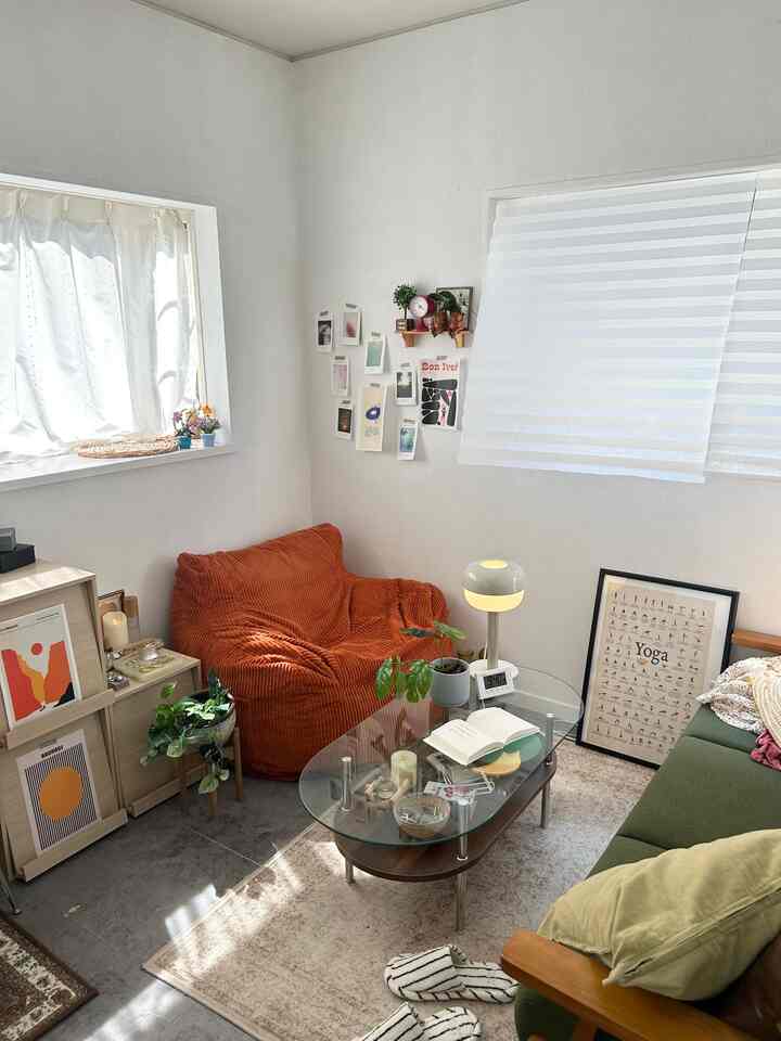 白い壁と大きなブラインド付き窓、オレンジのアームチェアとグリーンのソファが配置された雰囲気のあるナチュラルモダンのリビングルーム