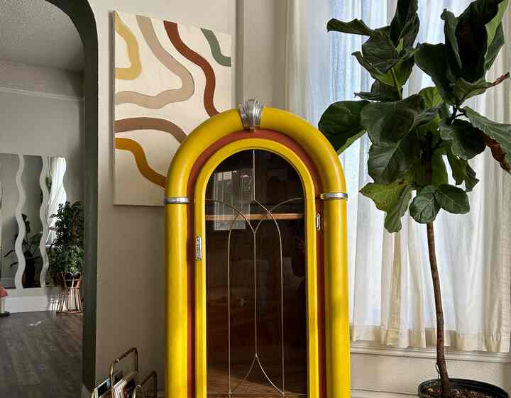 Cozy living room with beige-toned walls, vintage yellow jukebox cabinet, and a large plant near sheer curtains illuminated by natural light