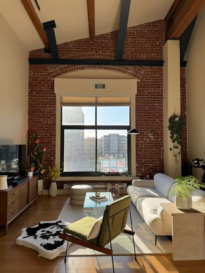 Mid-Century Modern living room with white sofa, green armchair, wood flooring, and exposed brick wall illuminated by natural light