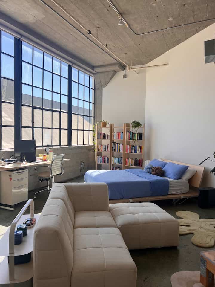 Bright white and natural wood toned compact bedroom home office featuring blue bedding and neat sofa in a minimalist space