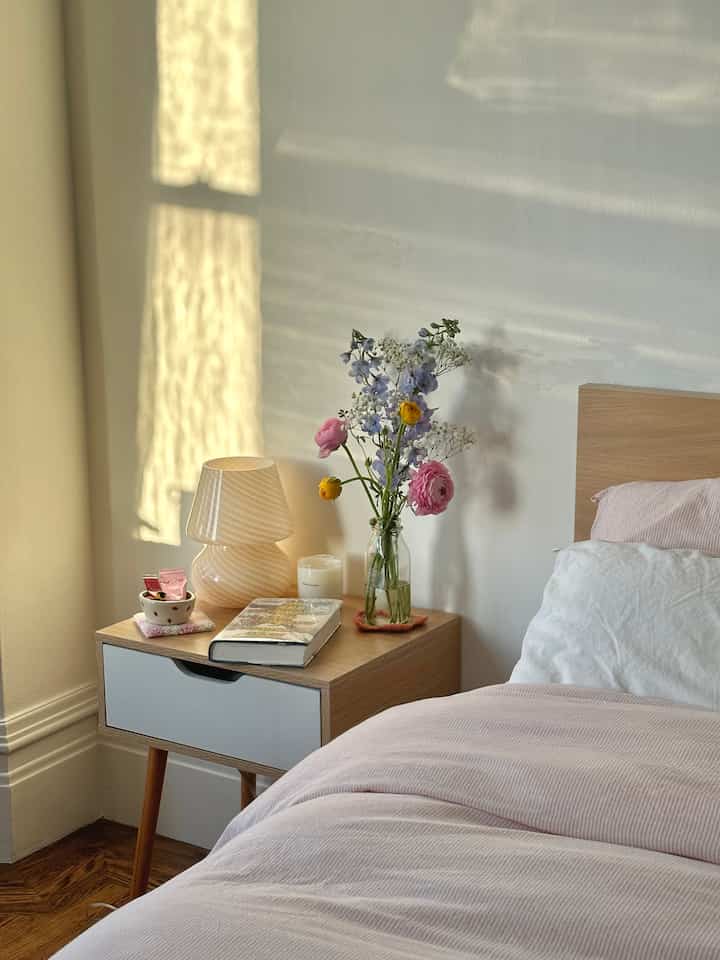 Natural toned bedroom featuring wooden bed, nightstand with vase and table lamp creating cozy atmosphere