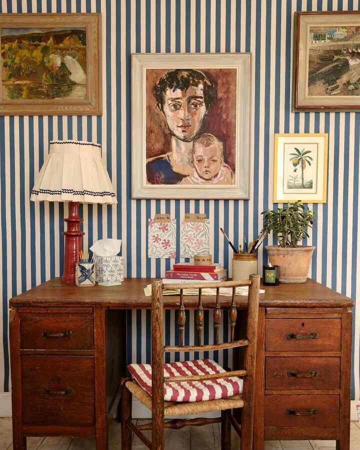 Blue and white striped wallpaper background featuring wooden vintage desk and chair, antique lamp and paintings creating a French country home office setting