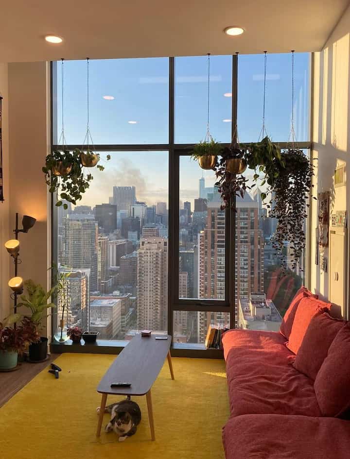Modern living room with a yellow rug, red sofa, and hanging planters featuring city skyline views through large windows