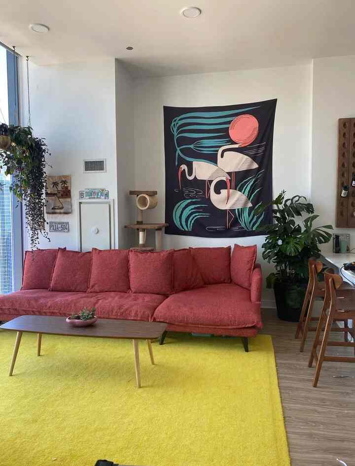 Living room with a yellow rug and red sectional sofa, featuring large plants and a cat tower in a natural setting