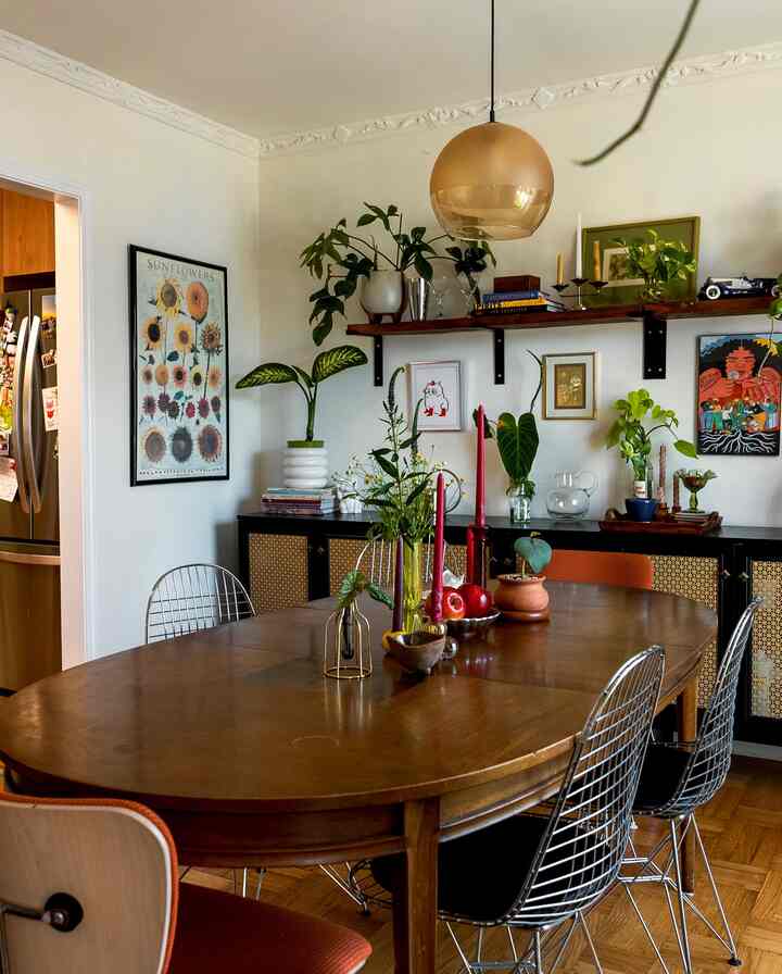 A natural brown and green toned dining room featuring a wooden dining table, wire chairs, plants on the table, and an amber pendant light creating a cozy atmosphere