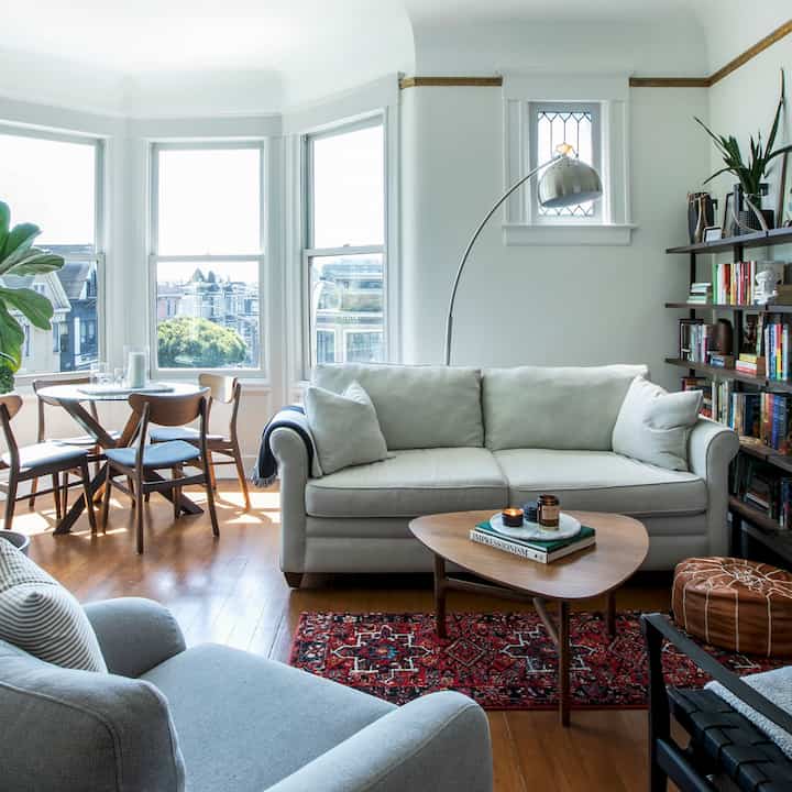 Bright natural toned living room featuring large windows, gray sofa, wooden tables, and bookshelf in a cozy atmosphere