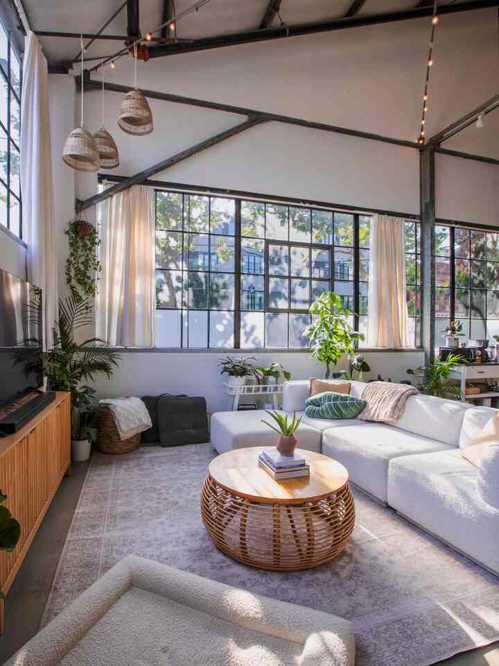Bright white and wood-toned loft living room featuring a large sectional sofa, round ottoman coffee table, and multiple plants for a natural atmosphere
