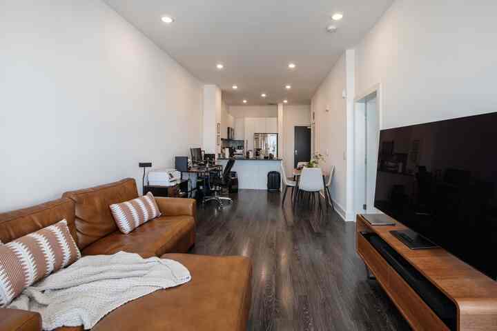 Bright white walls and ceiling in a long narrow room featuring a brown sofa, white dining chairs, and a modern living room combined with a home office