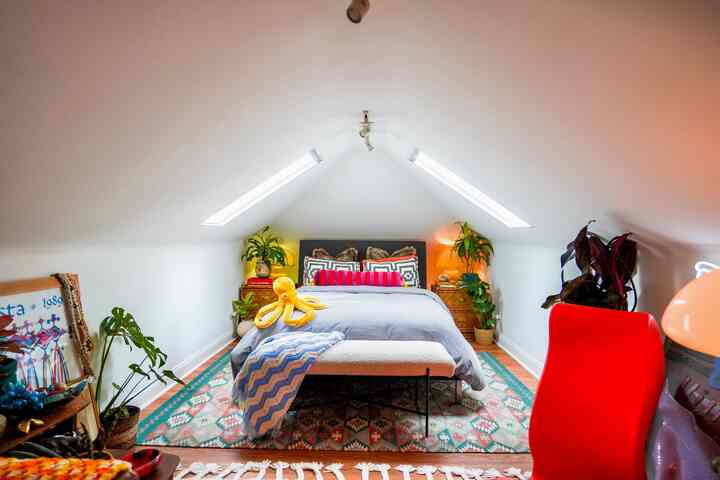 White-walled attic bedroom with slanted ceiling, featuring a central bed and bench surrounded by plants and table lamps creating a cozy atmosphere