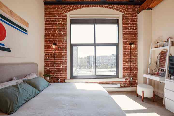 A bright mid-century modern bedroom with white walls and exposed red brick, featuring a bed with green pillows and wall sconces by the window.